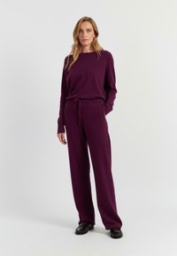 Paarse gebreide trui en bijpassende broek, met een ontspannen pasvorm, lange mouwen en een koordtaille, gecombineerd met zwarte schoenen.