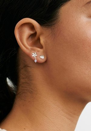 Naise kõrva lähivõte, kus on kaks hõbedast kõrvarõngast: väike õis rippuva helmestega ja lehekujuline kõrvarõngas.