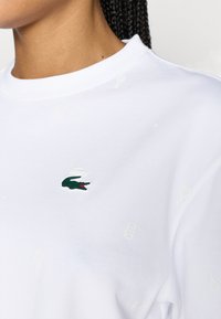 Λευκό βαμβακερό φούτερ με στρογγυλή λαιμόκοψη, διακοσμημένο με πράσινο κεντητό λογότυπο κροκόδειλου και δ ιακριτικά λευκά μοτίβα με θέμα το τένις.