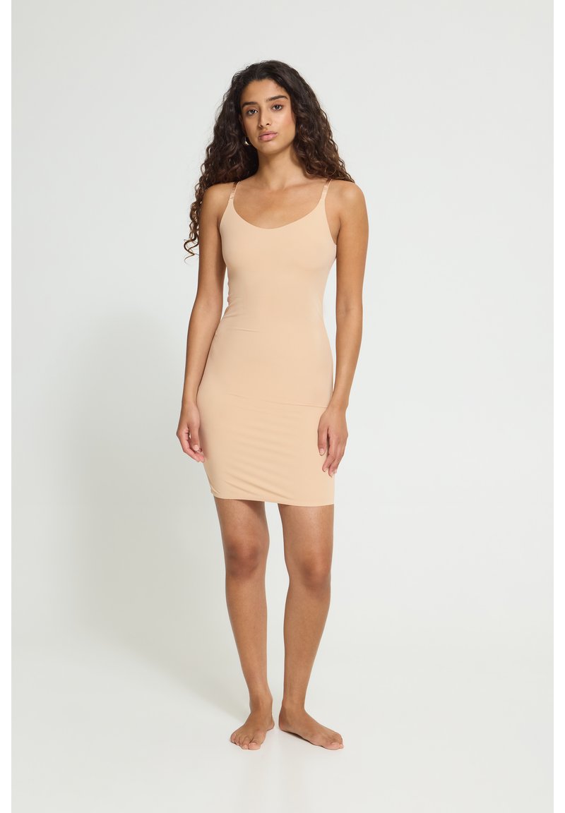Femme aux longs cheveux bouclés, debout pieds nus, portant une robe beige cintrée sans manches, longueur genou, sur un fond clair uni.