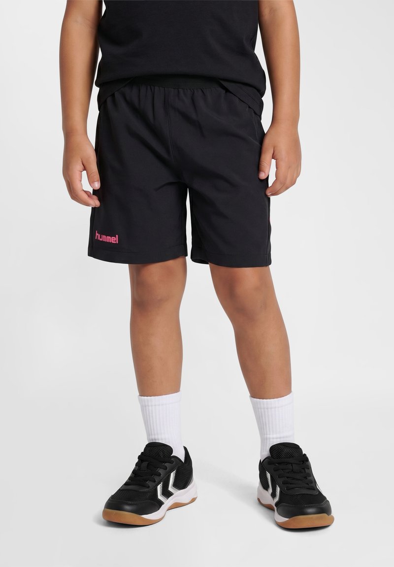 Dziecko noszące czarne krótkie spodenki sportowe z różowym logo, białe skarpetki do kostek i czarne buty sportowe stoi na jednolitym tle.