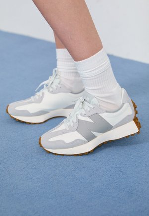 Baskets blanches et grises avec semelles en gomme, portées avec des chaussettes blanches montantes sur un sol recouvert d'un tapis bleu clair.