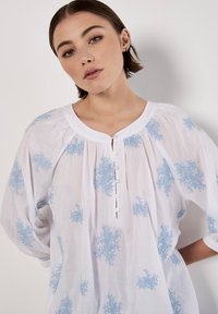 Vit blus med blå blomsterbroderi, rund halsringning, knappslå och vida trekvartslånga ärmar. Mjuk, lättviktig tyg.