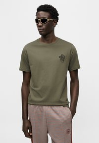 Πράσινο ελιάς βαμβακερό μπλουζάκι με μαύρο γραφικό ενός κάκτου σε γλάστρα στο αριστερό στήθος, σε συνδυασμό με καρό σορτς με σχέδια και μπεζ γυαλιά ηλίου.