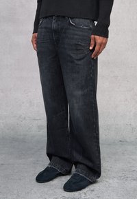 Jeans de mezclilla oscura con un corte relajado, que presentan una textura desgastada, costuras visibles y bolsillos laterales. Combinados con zapatos de tela oscuros.