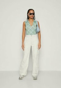 Πράσινη καρό αμάνικη μπλούζα, λευκά cargo παντελόνια και γκρι αθλητικά παπούτσια. Τα αξεσουάρ περιλαμβάνουν στρογγυλά γυαλιά ηλίου και μια μικρή μπλε τσάντα.