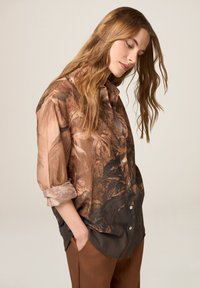 Camisa de botones con un patrón abstracto en marrón y beige, con mangas enrolladas y un corte holgado. Tejido suave con una textura blanda.