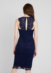 Vestido de encaje azul marino con una silueta ajustada. Presenta un corte en forma de lágrima en la espalda, bordes de encaje con forma de ola y un dobladillo a la altura de la rodilla.