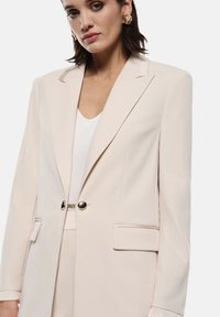 Blazer beige chiaro con una vestibilità su misura, rever a punta, due tasche frontali e bottoni neri. Tessuto liscio con silhouette ben definita.