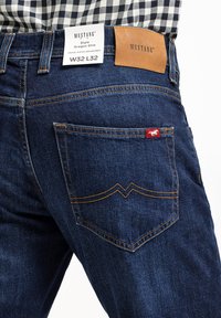 Dżinsy w ciemnoniebieskim kolorze z pomarańczowymi szwami, o dopasowanym kroju. Zawierają brązową skórzaną metkę z logo "MUSTANG" oraz małą czerwoną etykietę.