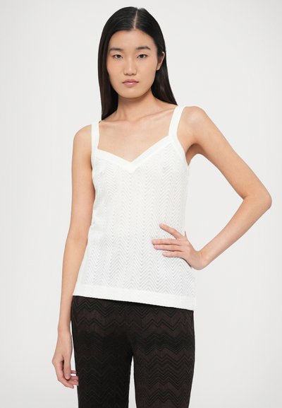 Λευκή αμάνικη μπλούζα με V-λαιμόκοψη και μοτίβο σε σχήμα βέλους. Οι τιράντες είναι φαρδιές και το ύφασμα φαίνεται ελαφρύ και ελαφρώς υφασμένο.