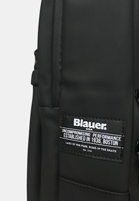 Zaino nero con una finitura liscia e opaca, dotato di scomparti con cerniera e un'etichetta in tessuto ben visibile con dettagli testuali e logo.