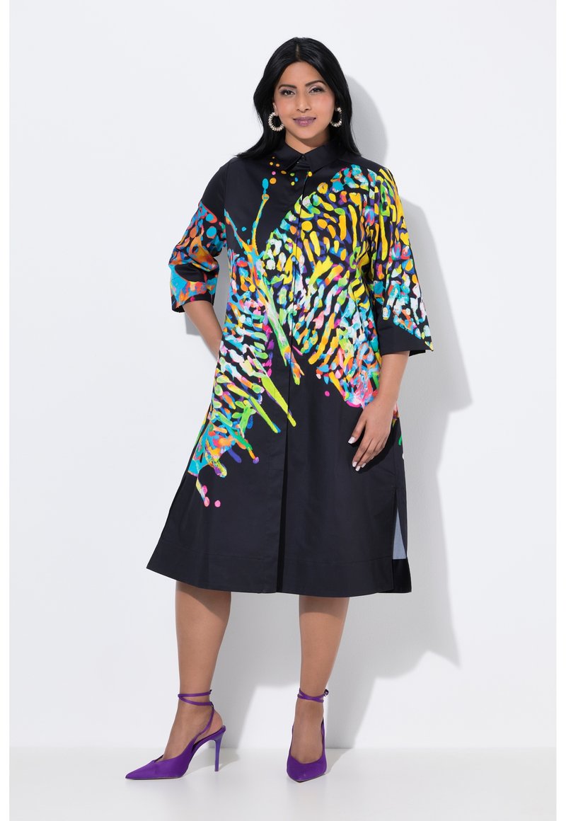 Schwarzes Kleid mit lebhaften, mehrfarbigen abstrakten Mustern, mit Kragen, dreiviertel Ärmeln und seitlichen Schlitzes. Kombiniert mit lila High-Heels.