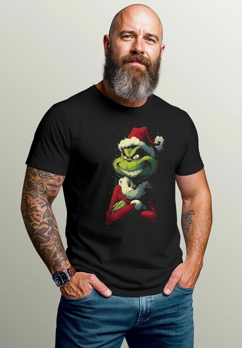 Bearded man with tattoos wearing a black T-shirt featuring the Grinch in a Santa hat, hands in pockets, standing against a plain background.