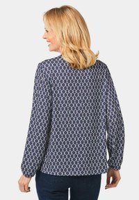Marineblauw blouse met een wit geometrisch patroon, lange mouwen, ronde halslijn en een plooitje aan de achterkant. De stof lijkt soepel en lichtgewicht.