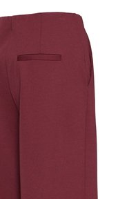 Gros plan d’un pantalon bordeaux montrant une poche arrière et une couture latérale avec une texture de tissu lisse sur un fond uni.
