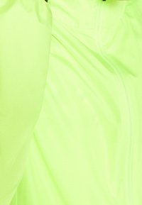 Veste légère vert néon avec une texture lisse, fermeture éclair et poignets élastiques. Le tissu semble résistant à l'eau.