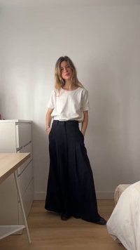 Kvinde med langt hår iført en hvid top med korte ærmer og højtaljede, sorte bukser med vide ben, stående i et minimalistisk soveværelse.