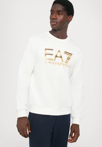 Sweatshirt branco de gola redonda com o logo dourado da EA7 Emporio Armani. Tecido macio com punhos e bainha canelados. Combinado com calças azul-marinho.