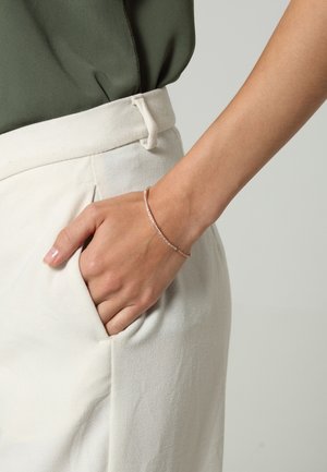 Main avec un bracelet fin en argent reposant à l'intérieur de la poche d'un pantalon à taille haute crème, associé à un haut vert olive.