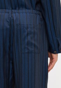 Blue striped jumpsuit featuring a herringbone pattern, elastic waist, side pocket, and buttoned cuff detail on the sleeve.