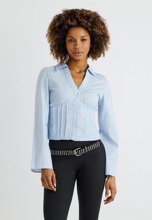 Kvinne med krøllete hår iført lys blå skjorte med tynne striper, knapper og vide ermer, samt svart belte med nagler på svarte bukser, nøytral bakgrunn.