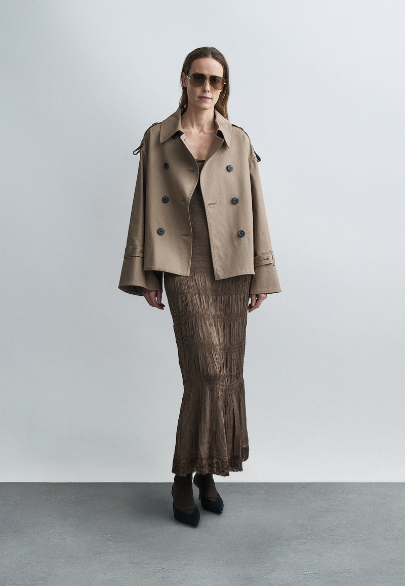Trench-coat beige à double boutonnage avec manches surdimensionnées, associé à une robe maxi plissée marron ajustée, dotée d'un ourlet évasé et d'une texture.