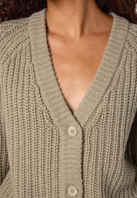 Close-up de uma pessoa a usar um cardigan de malha grossa em bege com decote em V e grandes botões, mostrando o pescoço e a parte superior do peito.