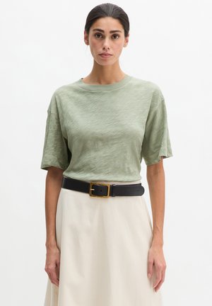 Femme aux cheveux foncés portant une chemise verte clair à manches courtes rentrée dans une jupe beige avec une ceinture noire à boucle dorée.