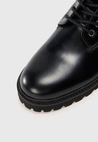 Botte en cuir noir avec une finition lisse, bout rond et semelle épaisse à traction. Dotée de lacets noirs et d'œillets en métal.