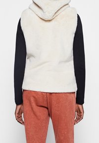 Krämfärgad, fluffig ärmlös väst med huva, med en mjuk textur och enkel design; bärs över en svart långärmad topp och rostfärgade byxor.