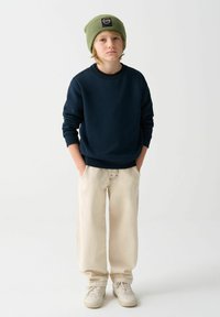 Námornícky sveter, béžové široké nohavice a krémové tenisky. Zelená pletená čiapka s logom. Neformálny outfit s uvoľneným strihom a textúrou.