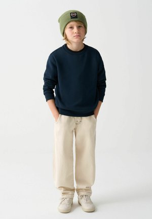 Námornícká mikina, béžové široké kalhoty a smetanové tenisky. Zelená pletená čepice s detailním logem. Neformální outfit s uvolněným střihem a texturou.