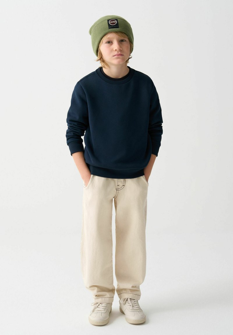 Námornícky sveter, béžové široké nohavice a krémové tenisky. Zelená pletená čiapka s logom. Neformálny outfit s uvoľneným strihom a textúrou.