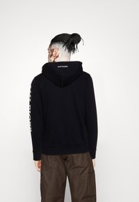 Haut à capuche noir avec une grande capuche, des poignets et un ourlet côtelés. Présente le logo 'HILFIGER' en blanc au dos et le long de la manche. Tissu doux et texturé.