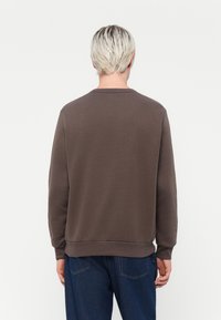 Brown sweatshirt with a crew neck, long sleeves, and ribbed cuffs; smooth texture and casual fit. Worn with dark blue jeans.