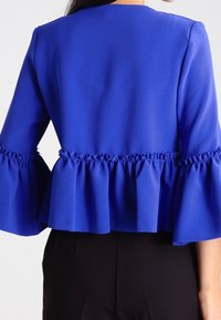 Haut bleu à basque volantée avec manches évasées, en tissu lisse. Présente des détails froncés à la taille et aux coutures des épaules.