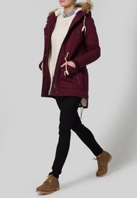 Burgundyröd parka-jacka med pälsfodrad huva, framfickor och dragsnören, bärs över en krämfärgad tröja och svarta byxor, parat med bruna stövlar.