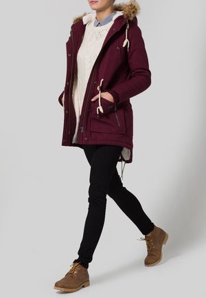 Parka burdeos con capucha forrada de pelo, bolsillos delanteros y cordones, llevada sobre un suéter crema y pantalones negros, combinada con botas marrones.