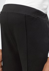 Persona che indossa pantaloni neri a vita alta e una camicia bianca, mostrata dalla vita in giù con vista laterale dell’anca e della mano.