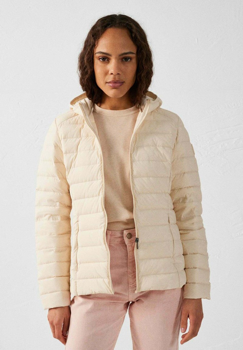 Beige donsjacket met horizontale stiknaden, voorzien van een capuchon en ritssluiting, gedragen over een lichtbeige top en lichtroze broek.