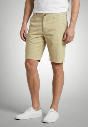 Mann trägt beige knielange Shorts, weiße Sneaker und ein weißes T-Shirt, steht vor einem einfarbigen grauen Hintergrund.