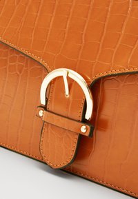 Brązowa torebka z eko skóry krokodyla z detalem w postaci złotego klamry, z kontrastującym przeszyciem i eleganckim, prostokątnym kształtem.