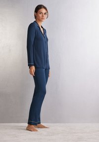 Mörkblå pyjamas set med en knäppskjorta och byxor med elastisk midja, med vit piping längs kragen och ärmsluten. Mjuk tyg.