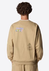 Beige sweatshirt med ribbad krage och muddar har färgglad text "EXPLORERS GATHER" och lekfulla illustrationer på baksidan och ärmarna.