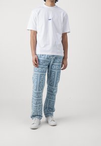 Camiseta blanca con un logo azul, combinada con jeans de un azul claro que presentan un patrón repetido de "HUGO". Terminada con zapatillas blancas.