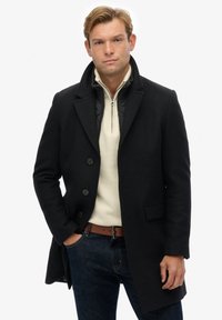 Abrigo de lana negro con solapas en pico, tres botones y bolsillos laterales; superpuesto a un suéter crema con cremallera y jeans oscuros con un cinturón marrón.