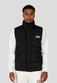 Gilet invernale nero con colletto alto, zip frontale e tasca sul petto con zip; presenta il logo sul lato sinistro. Indossato sopra una felpa bianca.