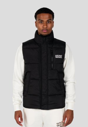 Black puffer vest with a high collar, front zipper, and zippered chest pocket; features logo on the left side. Worn over a white sweatshirt.