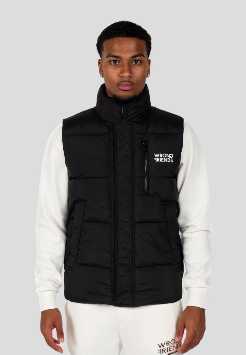 Gilet invernale nero con colletto alto, zip frontale e tasca sul petto con zip; presenta il logo sul lato sinistro. Indossato sopra una felpa bianca.